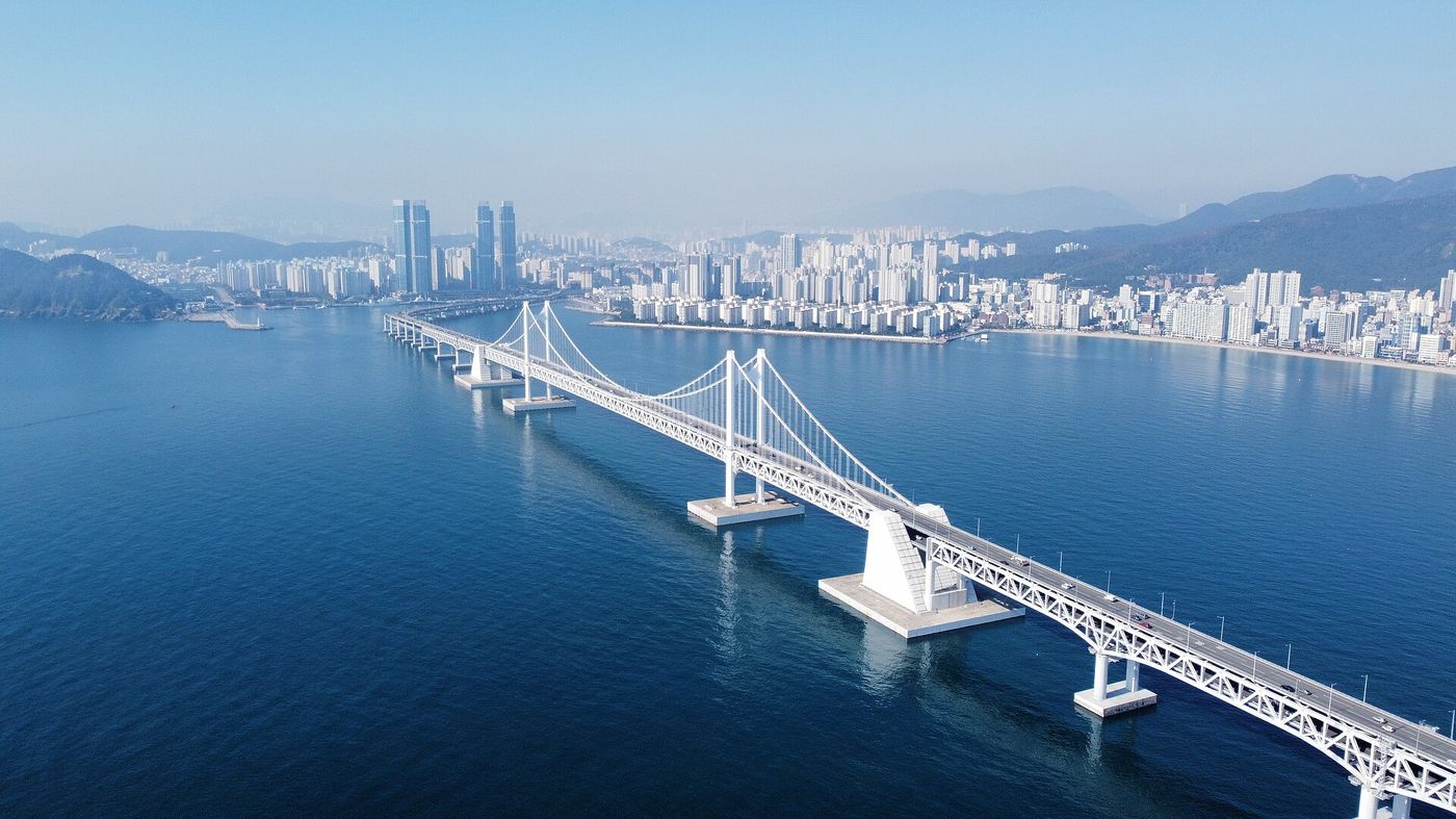 맑은 날 상공에서 내려다본 광안대교의 흰 현수교 전체와 부산 도심