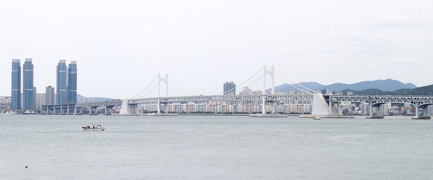 흐린 하늘 아래 원경으로 걸친 광안대교와 앞바다를 지나는 작은 어선 한 척