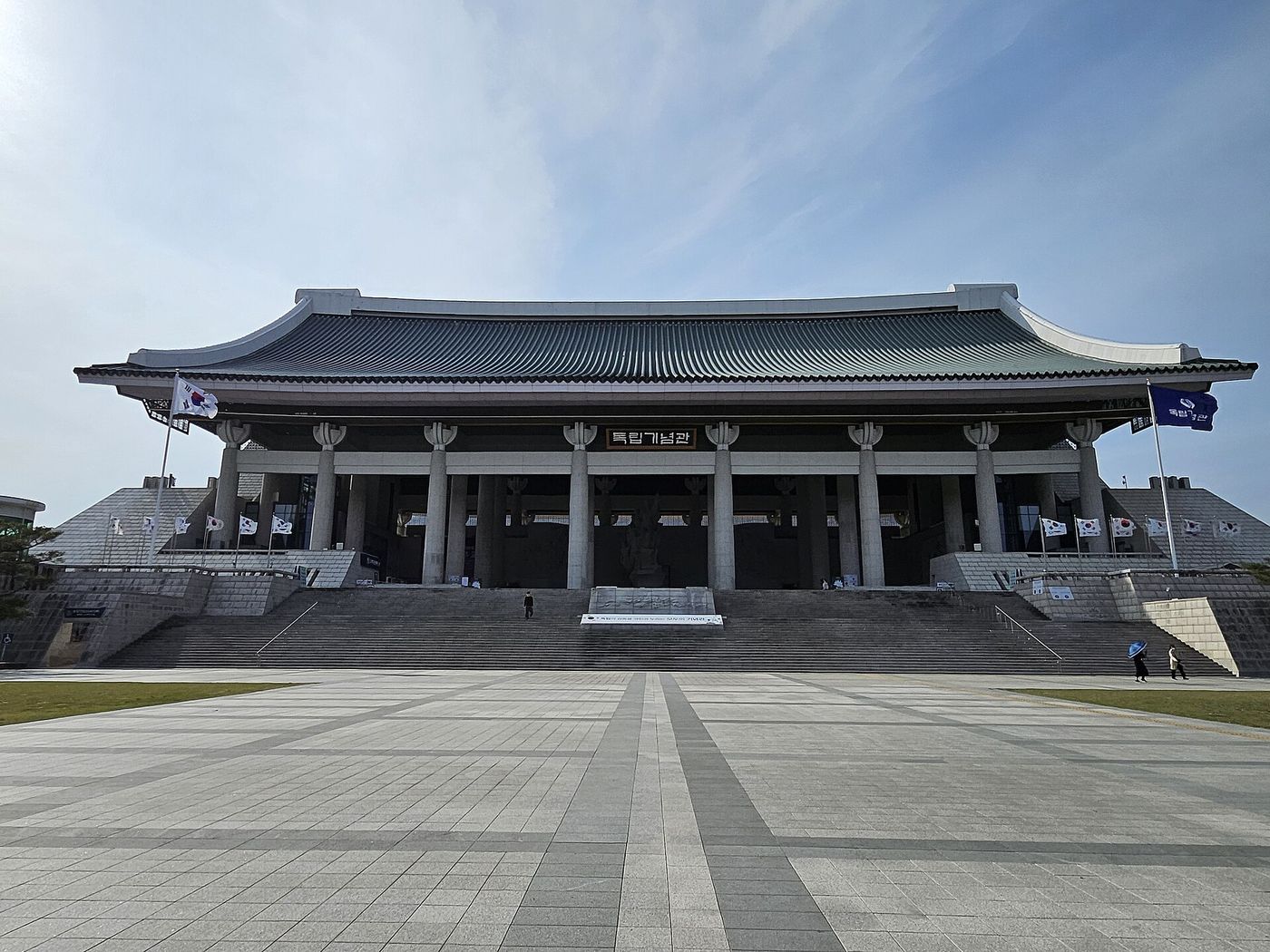 독립기념관 겨레의집 — 국내 최대급 기와지붕 아래 '독립기념관' 현판이 걸린 정면, 태극기가 늘어선 넓은 광장이 펼쳐진다