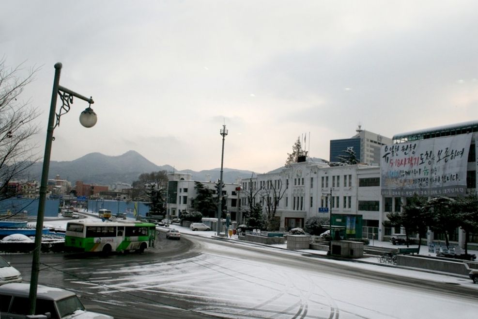 눈 덮인 옛 전남도청 앞 — 흰 건물 옆으로 '5·18사적지 도청을 보존하여…'라 적힌 현수막이 걸려 있다