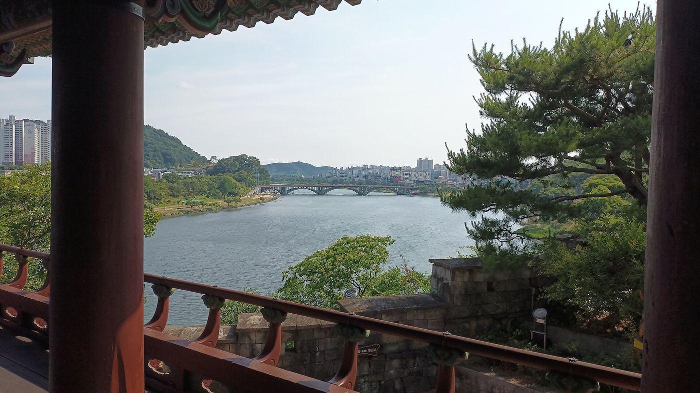 촉석루 마루에서 기둥 사이로 굽어본 남강 — 강 건너 도심과 강을 가로지르는 다리가 한눈에 드는 조망
